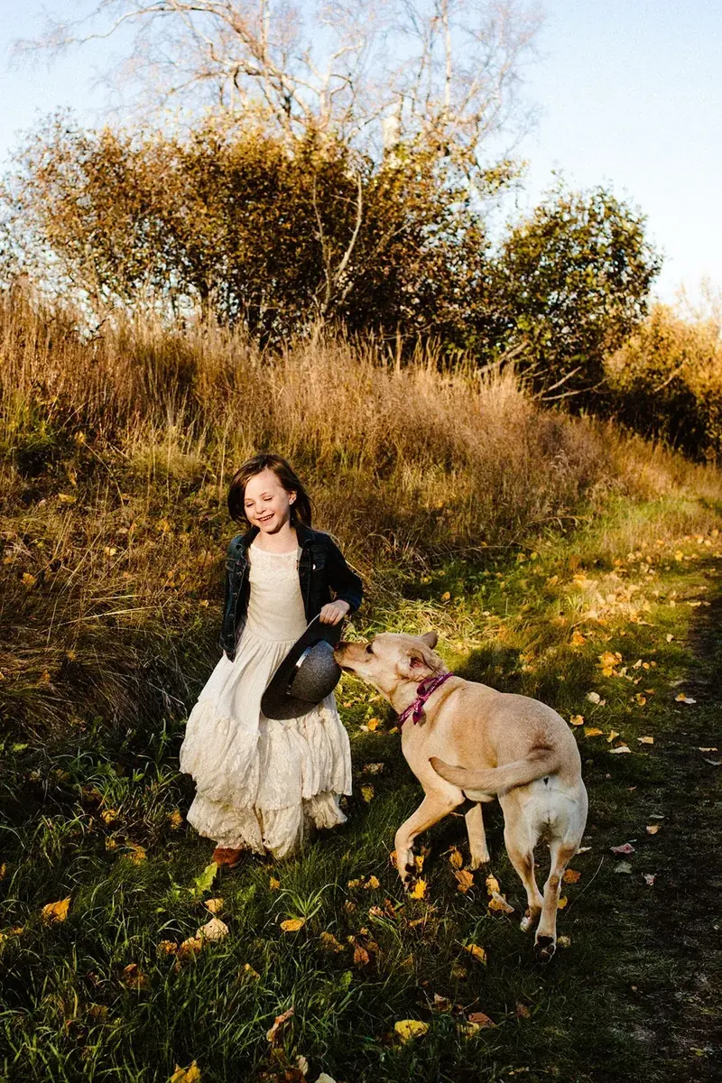 A laughing young girl in a white dress and denim jacket walking a yellow Labrador along an autumn trail